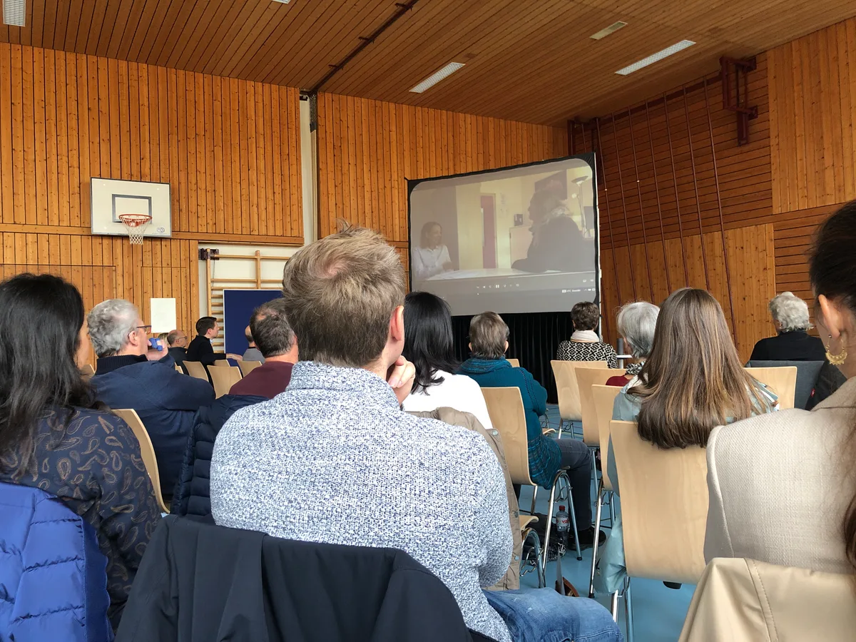 In einer Turnhalle sitzen die Versammelten vor einer Leinwand.
